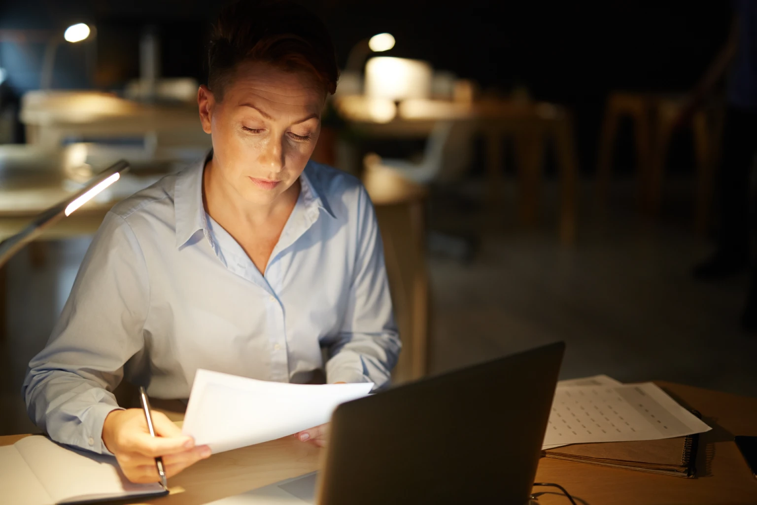Mujer analizando documentos en un escritorio iluminado por una lámpara, de noche